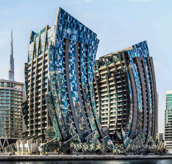 Modern architectural towers with fragmented glass facades overlooking a canal, with the Burj Khalifa in the background.