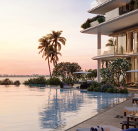 A resort-style swimming pool with lounge chairs and palm trees at sunset overlooking a large body of water.